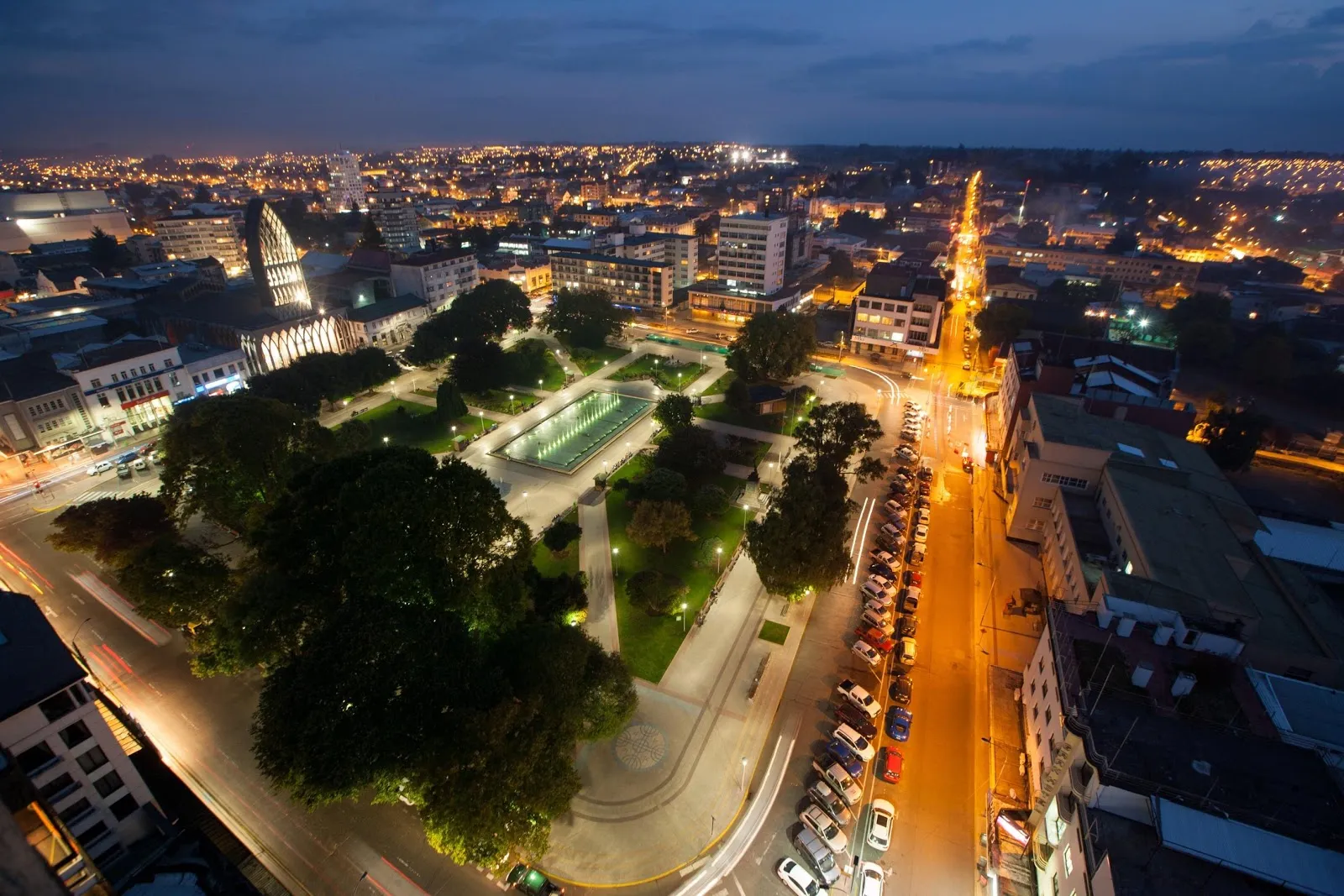 Foto aérea nocturna del centro de Osorno: la Plaza de Armas iluminada con una pileta rectangular y árboles, la catedral a un costado y una avenida con luces de autos y edificios alrededor.