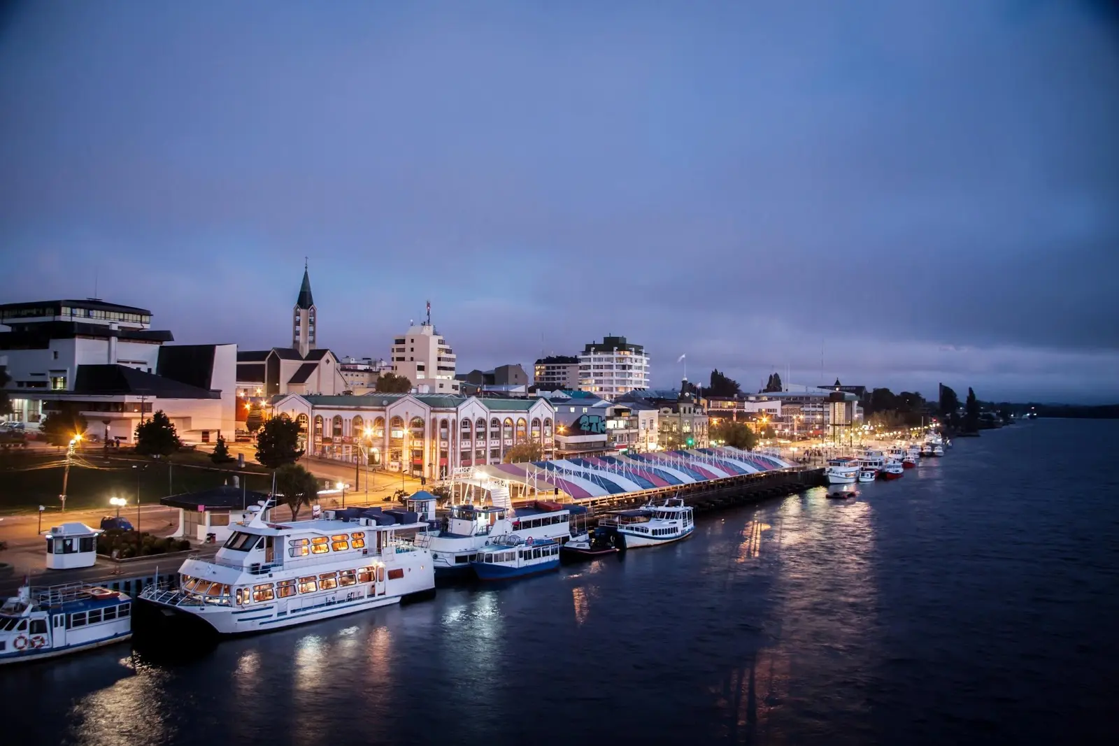 Costanera de valdivia  al atardecer con embarcaciones y luces reflejadas en el río.